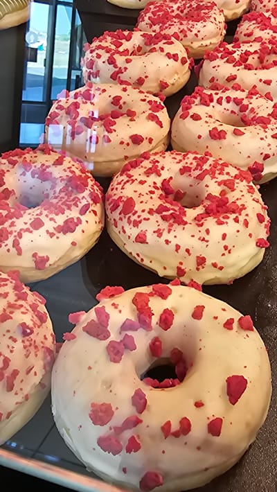 A close-up shot displays a collection of donuts with white icing and red sprinkles, arranged on a dark surface. The donuts are the primary subjects, and their round shape with a hole in the center is clearly visible. The white icing appears smooth and evenly distributed, while the red sprinkles, which are small and irregularly shaped, are scattered across the surface of each donut. Some of the sprinkles have a crumbly texture. The lighting is bright, highlighting the textures and colors of the donuts. The scene suggests a display of baked goods, possibly in a bakery or a food shop. There are no people visible in the image. The overall impression is one of sweetness and indulgence, with the vibrant red sprinkles adding a pop of color against the white icing. The context of Cuarte de Huerva, Spain, places this scene within a specific geographic location, but there are no overt visual cues in the image to confirm this location. There is no visible text in the image.