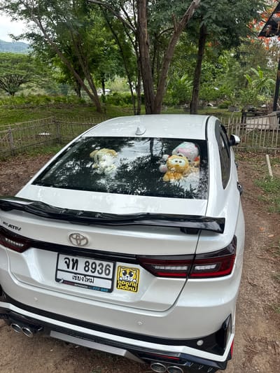 The image shows the back of a white Toyota Yaris Ativ sedan parked outdoors in Ban Huai Sai, Thailand. The license plate reads จท 8936 เชียงใหม่. Two stuffed animals are visible in the back window of the car. The car is parked on a dirt surface near trees and greenery. A metal fence is partially visible in the background.