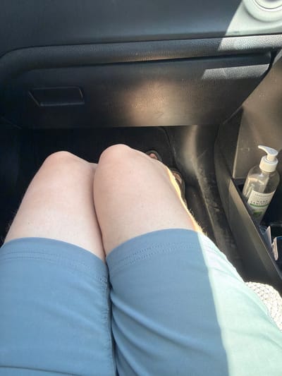 An overhead shot shows a person's bare legs in blue shorts, sitting in a car. Their feet, wearing sandals, are visible on the floor. To the right, a bottle of hand sanitizer sits in a cup holder. The car's dashboard is visible in the background. The scene takes place in Seaside, United States.