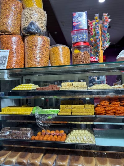 Here's a description of the image:
**Scene:** A street-side storefront in Kamalapura, India. The shop features a display of various snacks, sweets, and baked goods arranged on shelves behind a glass partition.
**Objects:**
* **Left side of display:**
* Packages of Indian "namkeen" (savory snacks), like sev, gathiya, and chivda, are stacked in plastic bags.
* Jars filled with peanuts and other small snacks
* Boxes of Parle Hide & Seek biscuits.
* Lollipops
* **Right side of display (sweets):**
* Jalebi (a sweet, fried dough).
* Burfi (Indian milk-based sweets) in multiple colors
* Laddu (ball-shaped sweets)
* Stacks of sliced bread.
**Environment:**
* The display is likely situated in a brightly lit shop, visible through the glass front.
* The storefront is likely in a busy commercial area, as evidenced by the variety of goods on offer.