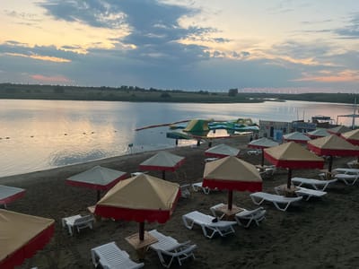 This image depicts a sandy beach area along a body of water, likely a lake, at dusk. The sky is filled with dramatic clouds painted with hues of orange, yellow, and blue, indicating the setting sun. On the water, there are inflatable water attractions, including slides and climbing structures. The beach itself is lined with numerous red and beige beach umbrellas, most of which are open, providing shade. White plastic lounge chairs are scattered across the sand, some arranged in rows and others individually placed. In the background, across the water, is a long, flat shoreline with sparse vegetation and a line of distant lights, possibly indicating a settlement. There are a few people visible on the beach and in the water, but they are not the primary focus of the scene. The overall impression is one of a recreational area at the end of the day. The location is identified as Байшуақ, Kazakhstan.