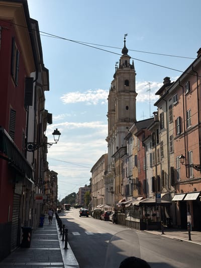This is a vertical, low-angle, outdoor photograph of a street scene in Parma, Italy. The image captures a view looking upwards towards buildings and a clear blue sky with some wispy clouds. The scene is oriented vertically in the frame, suggesting it might have been taken from a low perspective or that the photographer has rotated the image.
The subjects include several buildings lining a street, a church tower with a distinct spire and ornate architectural details, and a few people walking on the sidewalk. The buildings exhibit a traditional European architectural style with multi-story facades, numerous windows with shutters, and balconies. The church tower is a prominent feature, appearing to be made of stone with intricate carvings. On the street, there are vehicles, streetlights, and what appears to be a crosswalk.
The activity taking place seems to be everyday street life, with people going about their business. The time of day appears to be daytime, likely late morning or early afternoon, given the bright sunlight casting shadows. The weather is clear and sunny.
Notable details include the presence of electrical wires crisscrossing the sky, characteristic of older urban environments. The architecture suggests a historical setting, and the specific mention of Parma, Italy, provides a clear location context. The overall mood of the photograph is one of ordinary urban activity within a picturesque Italian city. There is no discernible text visible in the image.