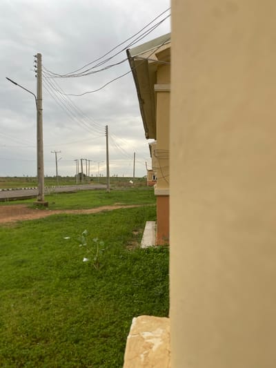 In Dumnga, Nigeria, a street scene unfolds under an overcast sky. Power lines crisscross the frame, supported by utility poles that stand along a paved road. The road curves gently to the left, bordered by a grassy median and a sidewalk. Adjacent to the road is a stretch of green grass, with a few scattered plants and some litter. In the foreground, a portion of a beige building occupies the right side of the image, partially obscuring the view. In the background, more utility poles and buildings can be seen stretching into the distance across a flat, grassy landscape dotted with sparse trees.