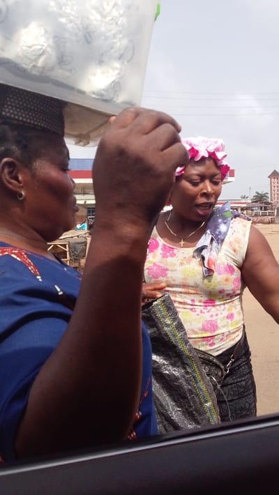 The image depicts an outdoor street scene in Igbo Ukwu, Nigeria, under an overcast sky. In the left foreground, a dark-skinned female, cropped from the chest up, wears a blue garment with a red and white pattern on the shoulder. A silver hoop earring is visible on her left ear. She balances a clear plastic container, appearing to hold foil-wrapped items, on her head, supported by a dark, mesh-like pad. Her right hand is raised near the container. Opposite her, in the midground, stands another dark-skinned female. She wears a pink ruffled head covering, a light-colored sleeveless top with a floral pattern, and a thin gold-colored chain with a cross pendant. A blue, white, and purple patterned fabric is draped over her left shoulder, and she wears dark, patterned trousers or a skirt. She holds a dark, woven bag with yellow and blue stripes. Her mouth is open, indicating speech. The two individuals are in close proximity and appear to be in conversation. The background reveals a dusty ground surface, a building with a red roof, a blue and red fuel station canopy, and a multi-tiered brown and white structure, consistent with a church, in the far right, alongside palm trees. A dark frame in the lower foreground suggests the photograph was taken from inside a vehicle.