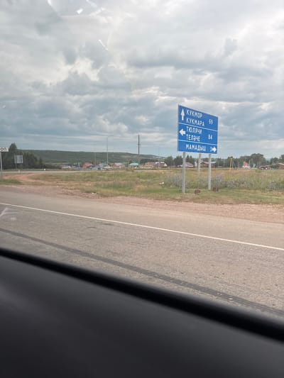 A blue directional road sign is visible against a cloudy sky. The sign points to "КУКМОР" (Kukmor) with a distance of 69, "КУКМАРА" (Kukmara) with a distance of 69, "ТЮЛЯЧИ" (Tyulyachi) with an arrow pointing left and a distance of 84, and "МАМАДЫШ" (Mamadysh) with an arrow pointing right and a distance of 3. The sign is positioned near a paved road with tire marks and a grassy area. In the background, a village with various houses and trees can be seen, with rolling hills in the distance. The location is identified as Красная Горка, Russia.