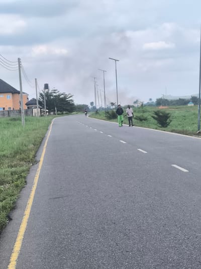 The image depicts a wide asphalt road stretching into the distance under an overcast sky in Obiaruku, Nigeria. A solid yellow line borders the left edge of the road, while white dashed lines delineate lanes further along. Lush green grass and low-lying vegetation line both sides of the roadway.

Two men are the primary subjects, walking side-by-side on the right side of the road, moving away from the camera. The man closer to the camera wears a dark jacket and bright, possibly neon green, trousers. His companion wears a light-colored shirt and dark trousers. A third, much more distant figure is faintly visible further down the road.

On the left side of the road, utility poles with power lines are prominent, standing in front of a partially visible two-story building with a reddish-orange facade and a dark roof. A black water tank on a stand is also visible near these structures. Modern streetlights with T-shaped tops line both sides of the road, extending into the background. In the mid-distance, a plume of light-colored smoke or haze rises above the horizon, visible behind the streetlights. The cloudy sky suggests an overcast day with soft, diffused lighting. No discernible text is visible.