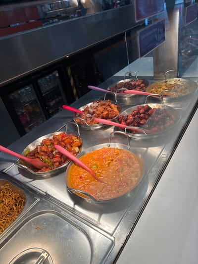 This image depicts a food service counter within a commercial establishment in the city of Dudley, United Kingdom. The scene is viewed through a transparent barrier, likely glass, showcasing multiple ready-to-serve hot food items presented in stainless steel buffet pans.
In the foreground, from left to right, a section of a metal well containing what appears to be noodles is visible. Adjacent to it is an oval-shaped pan holding stir-fried chicken pieces with green and red bell peppers in a reddish-brown sauce, containing a pink silicone serving spoon. Next, a larger oval pan displays two distinct liquid-based dishes, possibly curries or stews, separated down the middle, with a pink silicone serving spoon.
In the background, positioned on a second tier of the display, a row of five additional pans is visible. From left to right: a partially obscured pan containing a reddish-brown food item with a pink serving spoon; an oval pan filled with battered or fried chicken pieces coated in a light brown, glistening sauce and sprinkled with white sesame seeds, containing a pink silicone serving spoon; an oval pan holding dark reddish-brown chicken or meat pieces in a dark, glossy sauce, also with a pink silicone serving spoon; an oval pan containing lighter brown stir-fried food, possibly noodles or a vegetable mix, with a purple silicone serving spoon; and finally, a partially obscured pan on the far right containing a dark reddish-brown food item with a purple silicone serving spoon.
Behind the food display, a stainless steel counter reflects ambient lighting. Further back, a glass-fronted refrigerated display unit containing bottled beverages is visible. Overhead and to the left, additional metallic structures are present. No individuals are discernible within the frame.