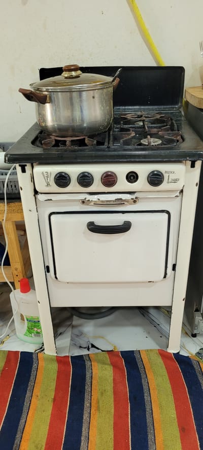 An old, white enamel gas stove with a pot on top is shown in a room in Zubrești, Moldova. The stove has four burners and a drawer below the controls. The pot is made of stainless steel with a glass lid and wooden handles. The stove is situated in front of a light-colored, textured wall. To the left of the stove, a bottle of green cleaning liquid is visible. A colorful striped rug covers the floor in front of the stove. The overall environment suggests a domestic setting, possibly a kitchen.