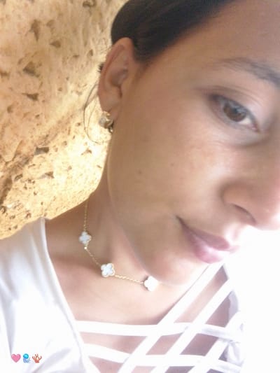 A close-up selfie shows a person with brown eyes and dark hair, wearing a gold necklace with white clover-shaped pendants and a white top with a crisscross design on the chest. The person is positioned in front of a textured, light-colored wall. The background wall is the city of El Cercado, Venezuela.