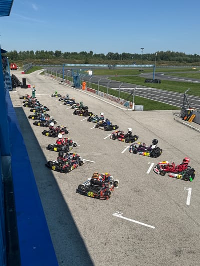 The image depicts a sunny day at a go-kart track, possibly located in Sekiotovo, Russia, with a clear blue sky and lush green trees lining the background. A row of go-karts, each with a driver in racing attire, is lined up on the asphalt track, suggesting an upcoming race or a pause during one. The go-karts are varied in color and design, with some displaying numbers and sponsor logos. A few individuals, likely participants or track staff, are visible near the starting line. The track features white lines marking lanes and a pit lane area. A blue barrier runs along the left side of the image, and a safety fence encloses the track. In the distance, more of the race track is visible, curving into the distance.