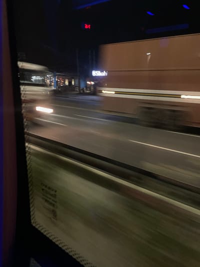 The image captures a night scene from what appears to be a moving vehicle, looking out a window. The perspective suggests a view of a highway or a well-lit road, possibly within or near the city of Samalkha, India, given the provided context. In the foreground, the window frame of the vehicle is visible, with some reflections and possibly condensation.

The main subject of the image is the road and the traffic. A large, brown truck with a trailer is prominent, moving from left to right across the frame. Its side has some reflective yellow and white stripes. The truck appears to be a commercial vehicle, possibly a goods transport truck. The road is asphalt, with clear white lane markings. Streaks of light indicate that other vehicles are also in motion, creating a sense of speed and blur.

In the background, there are some buildings and structures that appear to be shops or businesses lining the road. Some of these structures are illuminated by lights, and there are signs visible, though the text is largely illegible due to the distance and motion blur. One sign, partially visible and illuminated, contains the word "Shahi" in English. There are also some streetlights and possibly the lights of other shops in the distance. A red digital display, possibly a clock or a sign, is visible in the upper portion of the frame, displaying an indecipherable number or symbol. The overall atmosphere is that of a typical night scene on a busy road, with illuminated signs and moving vehicles. There are no clear individuals identifiable in the image, only the general impression of activity on the road and in the roadside establishments.