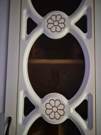 The image shows a close-up of a decorative panel, likely part of a door or screen, with an oval-shaped window in the center. The panel is white with raised, contoured edges and a gold trim. Two floral-like designs, also in white with gold outlines, are positioned above and below the oval window. The window itself is dark and appears to have a textured, possibly metallic, surface. The overall aesthetic suggests it might be a decorative element for a home or building. The location in the city of Қанабек, Kazakhstan is not discernible from the image content.