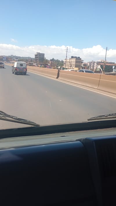 The image depicts a daytime scene from the interior of a vehicle looking forward through the windshield, specifically located in Kinoo ward, Kenya.
In the immediate foreground, the dark-colored dashboard and lower section of the windshield are visible, with two windshield wipers resting horizontally at the bottom of the glass. The midground shows a dry, paved multi-lane road, likely part of a dual carriageway. A light-colored pickup truck with a bed canopy, bearing a "KPM" series Kenyan license plate, is traveling in the left lane. Ahead of it, a yellow vehicle is partially visible. A light brown concrete barrier separates the visible road lanes from the right roadside.
The background reveals an urban environment. Beyond the concrete barrier, various buildings are present, including multi-story structures, some of which appear to be under construction with exposed frameworks. Electrical utility poles with power lines are spaced along the roadside. Patches of green vegetation are interspersed among the buildings and on distant elevated terrain. The sky above is predominantly clear blue with scattered white cumulus clouds.