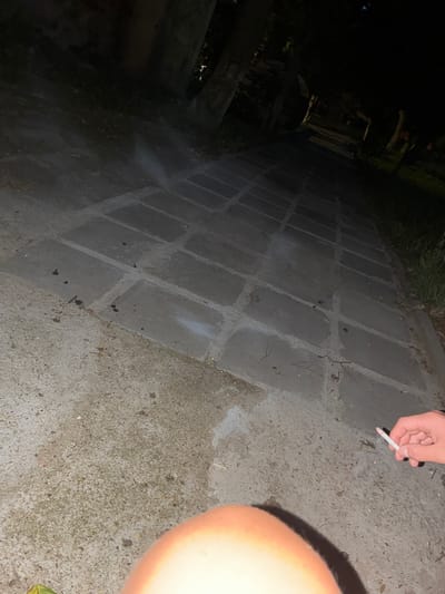 A close-up, low-angle shot captures a person's hand holding a lit cigarette in the foreground, with a blurred, light-colored rounded object, possibly a knee or thigh, also in the immediate foreground. The background depicts a paved pathway made of rectangular concrete slabs, leading into darkness. Trees and foliage are visible on the sides of the pathway, suggesting an outdoor, possibly park-like, setting in Glufishevo, Bulgaria. There is a faint wisp of smoke emanating from the cigarette. The lighting is dim, indicating it is nighttime or a poorly lit area. The overall scene is somber and suggests a moment of pause or contemplation.