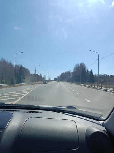The image shows a view from inside a car driving on a multi-lane road. The car's dashboard is visible in the foreground. The road is gray and has white lane markings. There are lampposts along the roadside. On either side of the road are trees, some bare and some with green foliage. The sky is blue with some haze or thin clouds. There are guardrails on one side of the road and a decorative fence on the other. A few cars are visible in the distance on the road. The general environment suggests a location in a forested area, potentially on the outskirts of a city or town. There are no visible people. The location may be near Kuz'mino, Russia, although specific details to confirm are not immediately apparent in the image.