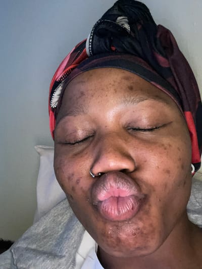 A close-up shot features a person with closed eyes, wearing a patterned headscarf. The person's face is turned towards the camera, with their lips pursed and a nose ring visible. The background appears to be an indoor setting, with a pale wall on the left and what looks like bedding or clothing in gray and white tones on the right. The lighting suggests it is daytime, but there are no specific weather cues. The overall mood is relaxed or contemplative, due to the closed eyes and facial expression. No text is visible in the image. The specific location within Randburg, South Africa, is not discernible from the visual information provided.
