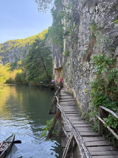 The image shows a view from the Iskar Panega Geopark near Petrevene, Bulgaria. A wooden walkway clings to a rocky cliff face. The walkway extends over a body of water, its surface reflecting the cliff and surrounding vegetation. People are seen on the walkway in the distance. In the foreground, a kayak with a paddle is partially visible. The sky is blue. The scene is dominated by the natural elements: rock, water, and vegetation.