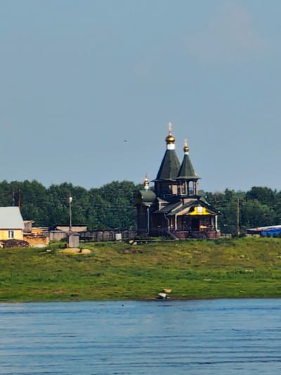 The image depicts a serene landscape with a prominent wooden church featuring green roofs and golden domes situated on a grassy hill. The church is surrounded by a few other buildings, including a yellow house with a white roof and what appears to be a small shed. A body of water, likely a river, occupies the foreground, reflecting the blue sky. In the background, a dense forest line stretches across the horizon under a clear, pale blue sky. A small dark object, possibly a bird or a drone, is visible in the sky. The overall scene suggests a peaceful rural setting in Потапово, Russia.