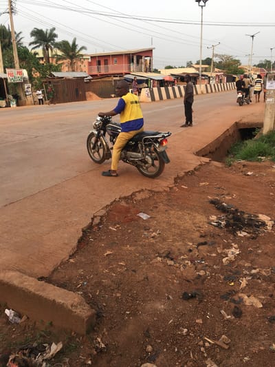 This image captures a daytime roadside scene in Isieke, Nigeria, likely in the late afternoon under an overcast sky. The photograph is oriented vertically.
In the foreground, a man wearing a yellow t-shirt with blue sleeves and yellow pants is riding a black motorcycle on a light-colored paved road. His t-shirt has partial text "EBRO..." visible. Further down the road, another man in dark attire stands near the roadside, and additional figures are discernible in the distance near buildings.
To the right of the paved road, there is a wide dirt shoulder and an embankment that is heavily littered with various types of rubbish, including plastic bags, paper, and organic debris. A concrete drainage channel runs parallel to the road.
The background reveals a mix of buildings, including a noticeable red multi-story structure, and smaller roadside stalls, some with corrugated metal roofs. Utility poles with extensive power lines crisscross the upper portion of the frame. Palm trees and other greenery are scattered throughout the setting.
Visible text includes "POS" on a white sign attached to a pole in the lower left, and "AT CALL" along with a partial phone number "08144000140" on a sign attached to a utility pole further down the road. The scene portrays a typical daily street activity in a bustling local environment.