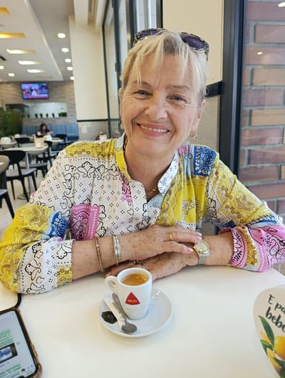 A woman is seated at a white table in an indoor cafe, possibly a coffee shop or restaurant, with a colorful, patterned shirt. She is looking at the camera and smiling. In front of her is a small white cup of espresso, a saucer, a spoon, and a packet of sugar. She is wearing a watch on her left wrist and several bracelets on her right wrist. She also appears to have a ring on her left hand. A smartphone is visible in the lower left corner of the image. The background shows other tables and chairs, and a television screen mounted on the wall, suggesting a public establishment. The decor includes exposed brickwork and modern lighting fixtures. The location is identified as Odivelas, Portugal, implying the cafe is situated there.