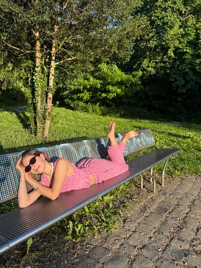 A young woman lies on a park bench in Hanau, Germany. She is wearing pink and white striped pants and a matching top, sunglasses, and a beaded necklace. She is barefoot and her hands are resting under her head. The bench is made of metal and has blue lettering on the slats. The ground next to the bench is paved with cobblestones. A grassy area with trees is visible in the background.