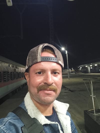 A man stands in Igatpuri, India, adjacent to a train carriage. He wears a camouflage cap, a denim jacket with a white, fluffy lining, and a dark shirt. A black shoulder strap is visible. The background shows a dark sky punctuated by streetlights, illuminating an expanse of pavement.