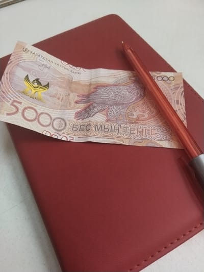 A 5000 Kazakh Tenge banknote is resting on a red leather-bound notebook. A red pen lies across the banknote. The scene appears to be indoors, on a white surface. There is no indication of the location being Töretam, Kazakhstan.