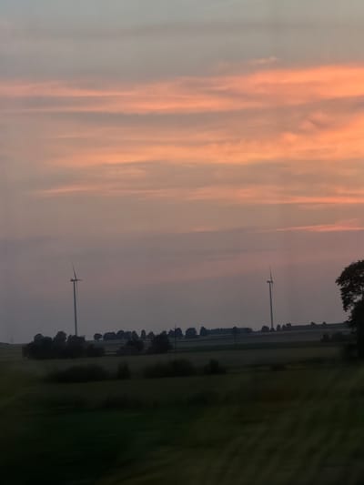 The image depicts a rural landscape at dusk. The sky is filled with bands of soft orange and pink clouds, characteristic of a sunset. In the middle ground, two wind turbines stand tall against the horizon, their blades silhouetted against the sky. A line of trees and vegetation forms a dark border below the turbines. The foreground is a blurred expanse of green fields, suggesting movement, possibly from a vehicle. The overall scene is serene and captures a moment of natural beauty in a rural setting. There are no people or discernible structures other than the wind turbines. The location is identified as Wola-Kolonia, Poland.
