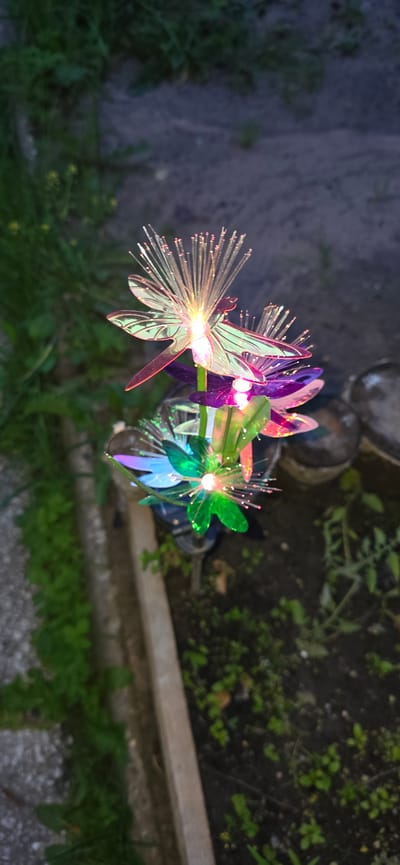 A close-up, vertical shot captures a decorative, illuminated garden ornament resembling a cluster of dragonfly-shaped flowers at dusk. The ornament is made of translucent, iridescent material with fiber optic strands emitting a soft, multi-colored glow. The colors range from pale pink and white at the top to green and blue at the bottom. The background consists of dark soil with scattered green foliage and a blurred concrete path on the left. The scene appears to be in an outdoor garden setting, possibly in Электротехник Снт, Russia, given the context provided. There are no people or animals visible in the image.
