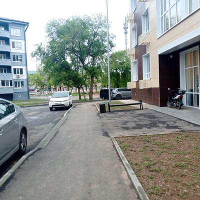 A sidewalk and parking area is shown in front of a modern building with beige and brown facade. A white SUV is parked on the street, and a silver sedan is visible on the left. A dark minivan and a silver SUV are parked further down the street. A small black scooter with a grey top box is parked near the entrance of the building. A metal bench and a trash can are located next to the sidewalk. Trees and other buildings are visible in the background. The scene appears to be in a city, likely Arseniev, Russia, given the context.