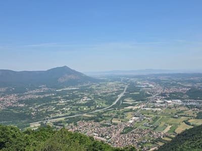 The image presents a panoramic vista over the city of Sant'Ambrogio di Torino, Italy, from an elevated vantage point. The scene is dominated by a clear blue sky. A mountain, possibly Monte Pirchiriano, sits in the mid-distance, its slopes covered in vegetation. Below the mountain and receding into the distance is the urban and agricultural landscape of the Po Valley.

A major highway cuts through the valley, connecting various towns and industrial zones. The city of Sant'Ambrogio di Torino is characterized by a dense cluster of buildings with predominantly red-tiled roofs. Agricultural fields, forests, and scattered buildings fill the spaces between towns and the city. In the foreground, lush green trees obscure the lower part of the image. There are no people visible in the image.