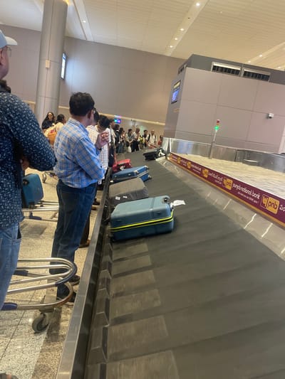The image shows a busy baggage claim area at an airport, likely in Gollapalle Khurd, India, as indicated by the Punjab National Bank advertisement prominently displayed on the baggage carousel. Several individuals are gathered around the carousel, waiting to collect their luggage. The carousel itself is moving, with various suitcases of different sizes and colors visible on it. Some people are looking towards the carousel, while others are standing or walking. The environment appears to be indoors, with a tiled floor and a ceiling with lighting fixtures. There are also pillars and structural elements characteristic of an airport terminal. The lighting is bright, suggesting it's daytime. The overall scene captures the common experience of air travel at baggage claim.