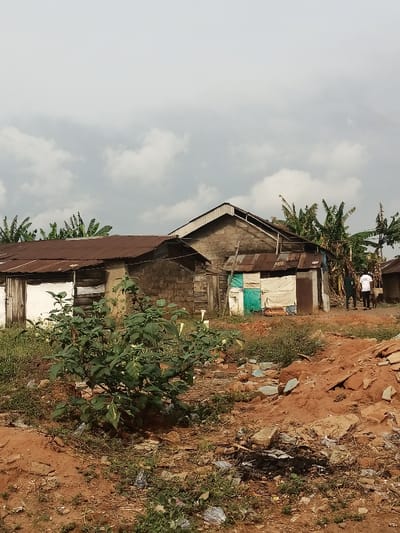 The image shows a partially built, two-story, concrete block structure with a corrugated metal roof in Obromudu, Nigeria, at the coordinates provided (latitude: 5.953, longitude: 5.697). A second structure with a corrugated metal roof, constructed of concrete blocks with partial wood framing, is partially visible in the image. The structures are located in a sparsely vegetated area and are situated on a patch of dry, red dirt with scattered debris. A large bush is in the foreground. Three people are walking on a path to the right of the structure, toward the back of the image. The sky is overcast with white clouds.