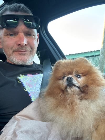 A man wearing sunglasses and a black t-shirt with a graphic print is in the driver's seat of a car. Next to him, a small, fluffy Pomeranian dog is nestled in a light-colored fabric carrier. The man is looking forward, while the dog is looking to the side. The background shows a clear sky and a hint of a green structure. This scene is taking place in the city of Kamyshovka, Russia.