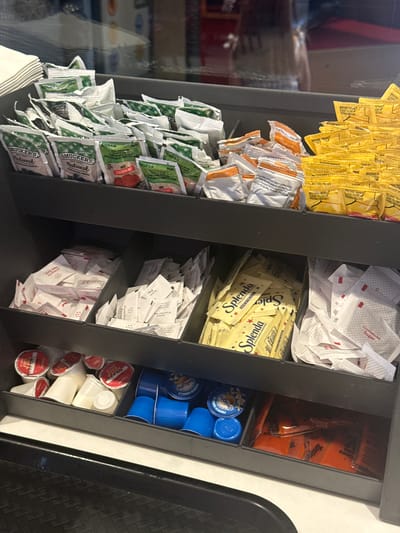 The image shows a condiment station, likely in a fast-food restaurant, possibly located in or near Mountain Park, Georgia, United States. The station is organized into several vertical sections. From left to right, the compartments contain: packets of yellow mustard, orange-labeled sauces, green packets of Smucker's Natural jam and other spreads. The middle section holds packets of white crystallized sugar, yellow Splenda artificial sweetener, and additional packets of white powder, possibly salt or pepper. The right-most section holds Tangy hot sauce, blue-lidded cups of creamer and half-and-half in individual containers. The station is made of dark gray material with a metallic accent. A black tray is visible on the right edge of the frame. The surroundings appear to be interior and somewhat dimly lit.