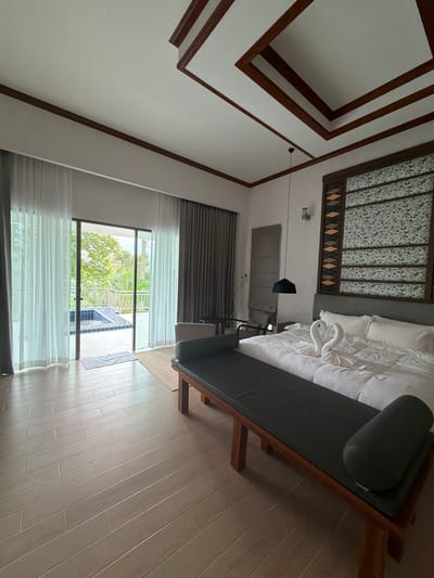 The image shows a bedroom with a king-size bed, a bench, and a door leading to an outdoor area with a small pool. The room has wooden floors and a ceiling with wooden trim. The bed is made with white linens and two swan-shaped towel arrangements. A dark bench with a cushioned seat is placed at the foot of the bed. The outdoor area visible through the door features a railing, lush green trees, and a blue-tiled pool. The overall impression is of a hotel room or a resort in a tropical location, possibly Ban Kao Dua, Thailand, given the natural greenery and the pool.