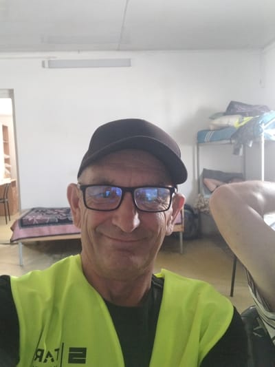 The media file is a portrait-style photograph, taken indoors, of a person wearing a bright yellow safety vest over dark clothing. The person has fair skin, short gray hair, and is wearing black-rimmed glasses and a dark cap. They are looking directly at the camera with a neutral expression. The background shows a room with what appears to be bunk beds, some bags, and a light fixture on the wall. The overall impression is of a casual, possibly utilitarian setting. The time of day is not discernible, nor is the weather. The location is stated as Oliivka, Ukraine. There is some text visible on the safety vest, but it is largely obscured and unreadable. The photograph is taken with a wide-angle lens, and the person is positioned slightly off-center to the left. The lighting in the room is somewhat dim, but the safety vest is brightly lit. The overall tone of the image is informal.