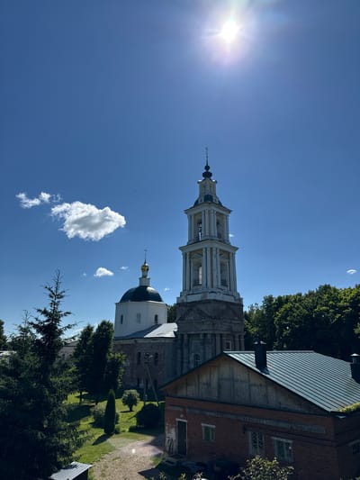 This is a photograph of the Bogoroditskaya Church in Vereya, Russia, on a sunny day. The image captures the church's prominent bell tower and a white dome with a golden cross. The bell tower is a tall, multi-tiered structure with arched openings. The church itself is partially visible, with white walls and a grey roof. In the foreground, there is a red brick building with a dark green metal roof. Surrounding the buildings are lush green trees and manicured lawns. The sky is a vibrant blue with a few scattered white clouds, and the sun is shining brightly, creating a lens flare in the upper right portion of the image. The overall scene depicts a serene and picturesque view of a historic religious site.