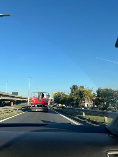 The image, taken from inside a vehicle, shows a view of a highway in Dock Sud, Argentina. A red VULCANO service truck is directly ahead. In the background, there are buildings and trees lining the roadside. A blue sky dominates the upper portion of the image. The image is rotated 90 degrees counterclockwise. The highway appears to be congested with traffic.