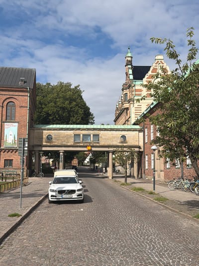 The image shows a street scene in Lund, Sweden, specifically the area around the Historical Museum of Northern Europe, which is part of the University of Lund.
In the foreground, a white Volvo car is parked on a cobblestone street. The car has the license plate "SJO 960". A light-colored trailer is attached to the car.
On the left side of the street, a brick building with a dark metal roof is visible. A banner with the text "MEDELTIDA KYRKLIG KONST HISTORISKA MUSEET" and an illustration of a church is hanging on this building. Next to it, a pedestrian bridge spans across the street, connecting two buildings. The bridge is made of stone and has arched openings and windows.
On the right side of the street, another brick building with white-framed windows is seen. A tree with green foliage is in front of this building. Several bicycles are parked along the sidewalk in front of the building.
In the background, more buildings with characteristic Scandinavian architecture, including a tower with a green copper spire, are visible. The sky is blue with scattered white clouds. The overall scene depicts a sunny day in a historic city center.