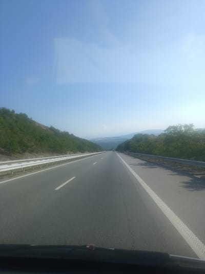 A highway view from a vehicle's perspective shows a clear blue sky with some reflective light streaks. The road ahead is asphalt with a white line marking the lane. Guardrails line both sides of the road, separating it from the surrounding landscape of green trees and hills. The scene appears to be on a highway, possibly near Blatino, Bulgaria, given the road infrastructure and the natural scenery.
