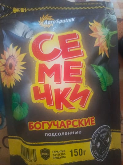 This is a close-up, slightly blurry photograph of the packaging for a product called "Семечки Богучарские" (Semechki Bogucharskie), which translates to "Boguchar Seeds." The packaging is predominantly black with large, bold red lettering that spells out "СЕМЕЧКИ" (SEMECHKI) in a stylized font with a yellow outline.
The packaging features illustrations of sunflowers on the left and right sides. On the left, a shaker of salt is depicted, suggesting the product is salted seeds. Below "СЕМЕЧКИ" and to the left, there are illustrations of green leaves.
At the bottom of the packaging, prominent text indicates "БОГУЧАРСКИЕ" (BOGUCHARSKIE) in red letters with a yellow outline, followed by "подсоленные" (salted) in smaller white text. To the left of this is a barcode, and the text "100% КАЧЕСТВА ПРОДУКТА" (100% QUALITY PRODUCT) in white and red on a yellow background. Next to this, the weight is clearly stated as "150 г" (150g).
On the right side, there's a circular emblem with Russian text that appears to be a quality seal, mentioning "ТОЛЬКО ОТБОРНЫЕ СЕМ" (ONLY SELECTED SEEDS) and "ЛУЧШИЙ СОРТ" (BEST SORT).
The "AgroSputnik" logo is visible at the top, alongside a zipper-like design element. The overall impression is of a commercial product packaging, likely for sunflower seeds. The lighting is somewhat uneven, and the background appears to be a textured fabric.