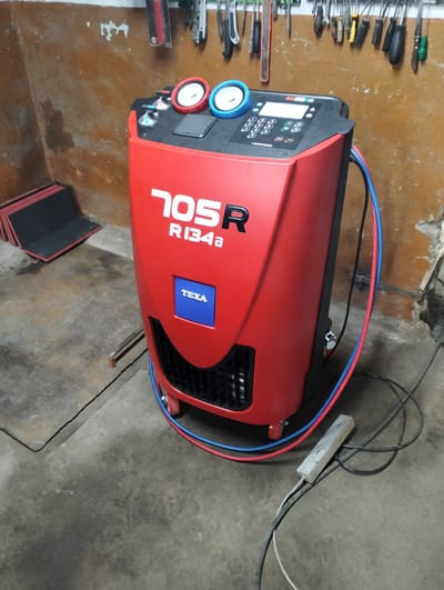 A red and black car AC service machine, identified as a TEXA 705R R134a, is positioned on a concrete floor. The machine has two gauges, one red and one blue, and a digital display with a keypad. Red and blue hoses are connected to the machine. In the background, tools are hanging on a pegboard against a rough, brown wall. To the left of the machine, a stack of black and red mats is visible. The setting appears to be a workshop or garage. The image is well-lit, suggesting it was taken indoors during daylight hours. No people are present in the image.