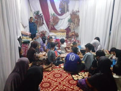 This image captures an indoor communal gathering, likely a celebratory event, in Gampongjawa, Indonesia. The setting features a decorated background with white and maroon draped fabrics, accented with artificial flowers and foliage, suggesting a wedding reception, engagement party, or a similar ceremony. A central platform or table adorned with fabric and holding two round pillows and a small white item serves as a focal point.
A diverse group of people, including adults and numerous children, are seated on a red patterned carpet covering the floor. Many women are wearing hijabs, and the attire generally appears modest. On the far left, a man stands next to a stack of folded textiles. The seated individuals are interacting, with some looking towards the center of the gathering. Scattered items, possibly food, drinks, or small gifts, are visible on the carpet among the people. One child in a blue jersey has the number "10" clearly visible on their back, and possibly the name "MUSLIM" (partially obscured) above it.
The lighting suggests it is either evening or an indoor event relying on artificial light. The atmosphere appears relaxed and communal, indicative of a family or community event.