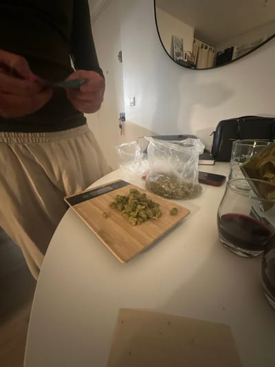 A person is shown sitting at a white round table. A scale with loose marijuana buds is on the table. A bag containing more buds is next to the scale. A large glass carafe, containing a dark liquid, is next to the bag. A black purse is in the background and a mirror is on the wall. The person has dark hair and is wearing beige pants and a long-sleeved dark green shirt. They are using a tool to manipulate the buds.