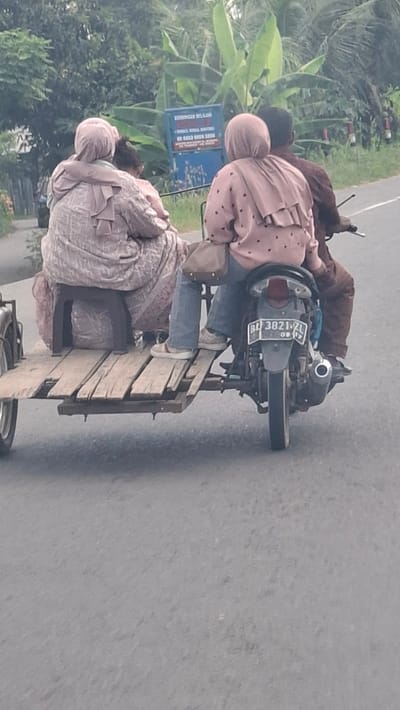 The image depicts a side view of a motorcycle with a sidecar attached, carrying three people on a paved road in Gampongrayadagang, Indonesia. The motorcycle is ridden by a person wearing a brown shirt, facing away from the camera. Seated in front of the rider is a person wearing a pink long-sleeved shirt with dark polka dots and jeans, also facing away from the camera. In the sidecar, a person wearing a patterned pink headscarf and a light-colored, intricately patterned dress is seated. This person is also facing away from the camera. The sidecar is constructed from wooden planks and appears to be a makeshift addition to the motorcycle. The environment includes lush green foliage and trees on the right side of the road, with a blue sign in the background. The road surface is gray asphalt.