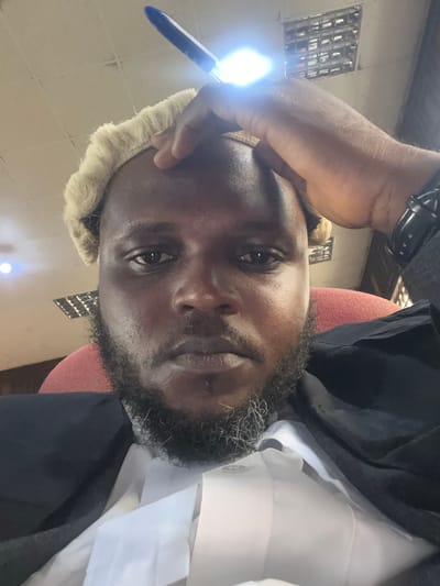 A close-up, slightly upward-angled selfie features a man with dark skin and a beard, positioned indoors in what appears to be a legal setting in Abeere, Nigeria. He wears a white collared shirt and a dark jacket or robe, along with a light-colored, possibly white or cream, legal wig. His expression is neutral and contemplative.
His left hand is raised, holding an object that emits a bright, intense light, possibly a pen with a built-in flashlight, positioned near his temple. A dark watch is visible on his left wrist. Behind him, a partial view of a red or maroon chair back is seen. The background includes a light-colored ceiling with visible paneling or a grid structure. To the left, a window with metal security bars or a distinct grid pattern is visible. Another diffuse light source appears in the lower right of the frame. The attire and setting strongly suggest he is a legal professional in a courtroom or office. No text is visible in the image.