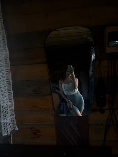 A woman is taking a selfie in a mirror. She is wearing a white crop top with a graphic print of mushrooms and the moon, and gray biker shorts. She has long brown hair and is wearing a watch on her left wrist. The mirror is in a room with wooden walls and a white lace curtain on the left. There are some clothes hanging in the background, and a wooden shelf on the right. The overall impression is that of a rustic, possibly rural, setting. The image is taken from a slightly low angle.