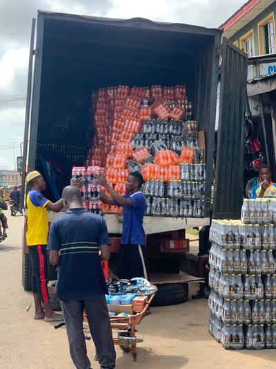 The scene is in Ondo, Nigeria, at the coordinates 7.251 latitude and 5.196 longitude. Three men stand near a delivery truck. The truck bed is full of cases of bottled beverages. One man is loading the truck with a case of bottled beverages. One man is standing near the truck with his hands on his hips. The third man is facing the camera and appears to be waiting. The truck has a large, green tarp on the side. The road is paved and there are other vehicles in the distance. There is a wheelbarrow with various items in the scene. The background is a typical African city with buildings and shops.