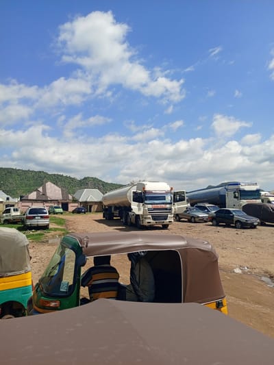 This media file depicts an outdoor scene in Barakin Babele, Nigeria, under a bright, blue sky with scattered white clouds. The time of day appears to be daytime, likely late morning or early afternoon, given the lighting. The setting is a dusty, open area, possibly a transport hub or a roadside stop, with several vehicles present.
The most prominent objects are a large white tanker truck with "MUBARAK IDEAL LTD" written on its side, and a smaller white truck. Other vehicles visible include several cars, a silver minivan, and two three-wheeled auto-rickshaws, one green and yellow, and another partially visible in the foreground. A person wearing a striped shirt is seated in the green and yellow auto-rickshaw. In the background, there are buildings with distinctive rooflines and a verdant, hilly landscape.
The activity appears to be the waiting or parking of vehicles, suggesting a moment of pause in transit. There are no overt signs of active work or movement, but the presence of multiple vehicles implies a place of some commercial or logistical significance.
Notable details include the textures of the dusty ground and the varied building structures. The weather is clear and sunny. The overall impression is one of a rural or semi-urban transportation setting in Nigeria.